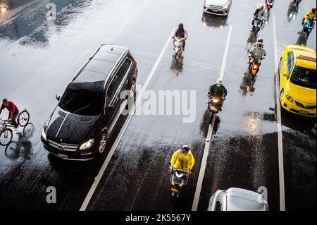 Autos, Minibusse, Busse und Motorräder fahren alle entlang einer belebten Kreuzung an einem regnerischen, dunklen Tag während der Hauptverkehrszeit in Bangkok, Thailand. Stockfoto