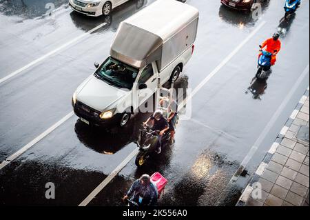 Autos, Minibusse, Busse und Motorräder fahren alle entlang einer belebten Kreuzung an einem regnerischen, dunklen Tag während der Hauptverkehrszeit in Bangkok, Thailand. Stockfoto