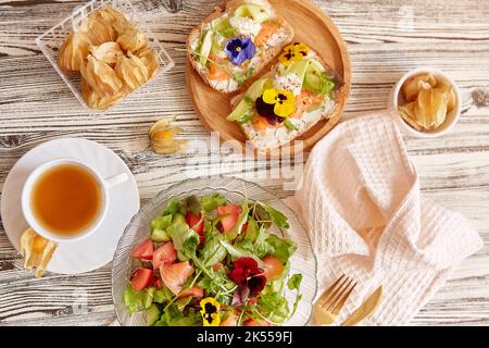 Ästhetischer Pescetarian-Tisch - Toast mit Lachs, Quark, essbaren Blumen. Mediterraner Salat mit frischem Gemüse und Lachs. Tasse Tee. Bei Stockfoto