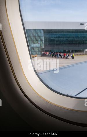 Blick von einem Flugzeugfenster auf einen Flughafen, mit einer Schlange von Menschen in der Ferne, die ihre Abflüge für ihr Flugzeug verlassen. Stockfoto