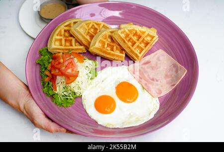 Hände halten Teller mit Eiern mit Schinken und Salat und belgischen Waffeln aus der Nähe Stockfoto