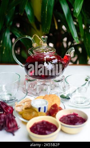 Hibiskus-Tee in einer Glas-Teekannen mit Tassen und kristallisierten Früchten und Marmelade auf einem Tisch Stockfoto