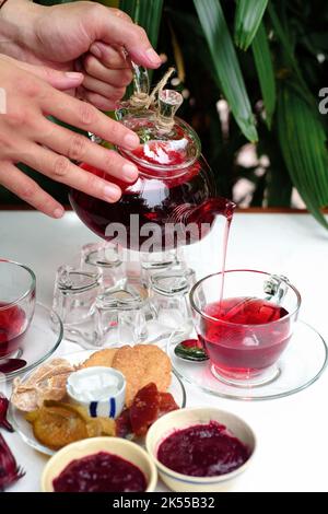 Hände halten Glas Teekannen mit Hibiskustee mit Tassen und kristallisierten Früchten und Marmelade auf einem Tisch Stockfoto