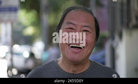 Glücklicher asiatischer amerikanischer Senior lächelt beim Gehen im Freien vor der Kamera. Aufgeregte Japaner mittleren Alters laufen auf dem städtischen Straßensteig Stockfoto