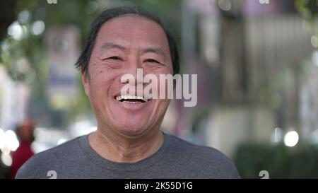 Glücklicher asiatischer amerikanischer Senior lächelt beim Gehen im Freien vor der Kamera. Aufgeregte Japaner mittleren Alters laufen auf dem städtischen Straßensteig Stockfoto
