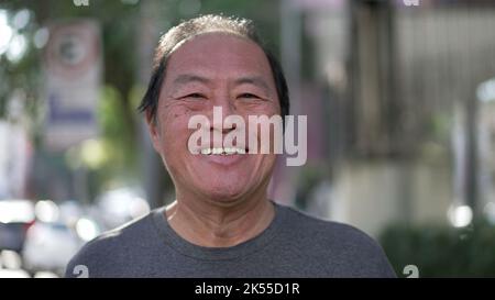 Glücklicher asiatischer amerikanischer Senior lächelt beim Gehen im Freien vor der Kamera. Aufgeregte Japaner mittleren Alters laufen auf dem städtischen Straßensteig Stockfoto