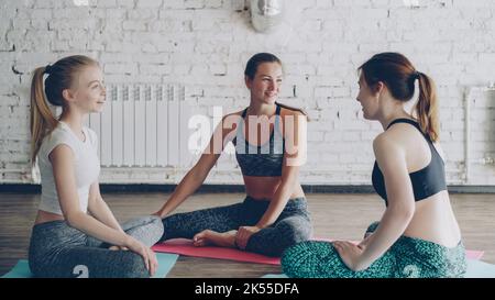 Junge, fröhliche, attraktive Frauen geselligen sich nach dem Yoga-Üben auf Matten im Kreis im Lichtstudio. Sie reden emotional, lachen, gestikulieren. Wellness Fun Konzept. Stockfoto