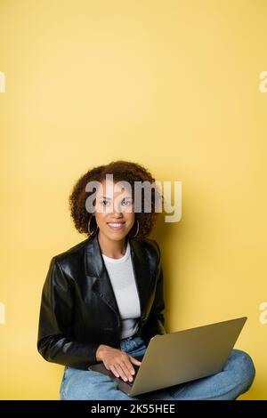 Fröhliche und junge afroamerikanische Frau in Lederjacke mit Laptop auf gelb, Stockbild Stockfoto