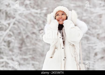 Weibliches Winterportrait. Schöne Frau in weißem Strickhut und warmer Kleidung, Jacke mit Fell bedeckt ihre Ohren mit Händen und lächelt im Frostschnee n Stockfoto