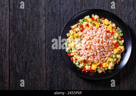 Gekochte kleine rosafarbene Garnelen auf Hirse-Pilaw-grünen Erbsen, roten Paprika und Mangostückchen auf schwarzem Teller auf dunklem Tisch, horizontale Ansicht von oben, Stockfoto