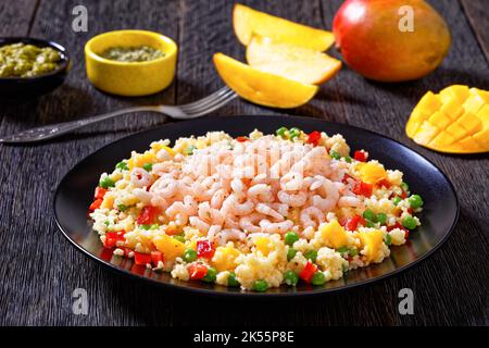 Garnelen überbacken Hirseporrige mit Gemüse, Mangostückchen und Gewürzen auf schwarzem Teller auf dunklem Tisch mit Zutaten im Hintergrund Stockfoto