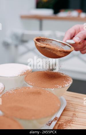 Der Prozess der Vorbereitung Tiramisu Dessert. Die Schale wird durch ein Sieb mit Kakao bestreut. Nahaufnahme, selektiver Fokus. Stockfoto