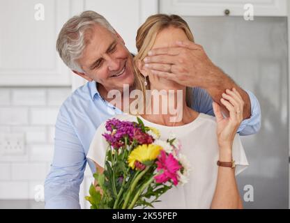 Seniorehepaar, das die Augen für Blumenüberraschung, Jubiläumsliebe und Valentinstag in Neuseeland zu Hause bedeckt. Glücklicher Mann, der der Frau für Blumenstrauß gibt Stockfoto