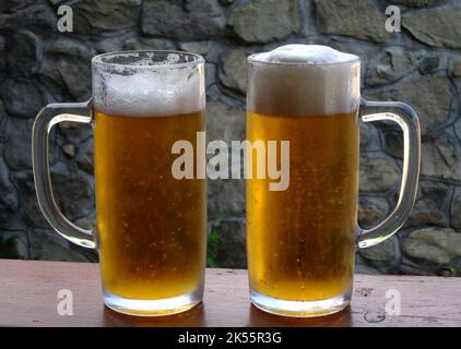 Zwei Gläser kaltes Bier vom Fass mit üppigem Schaum auf einem Holzschreibtisch mit Steinwänden Stockfoto