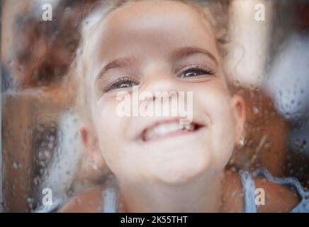 Fröhliches lustiges Mädchen, Gesicht am Fenster mit flacher Nase auf regnerischem Glas und Comic-Porträt zu Hause. Junges Kind spielen Witz, Kind allein mit großem Lächeln und Stockfoto