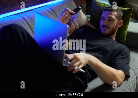 Fröhlicher, hübscher Kaukasusmann, der auf dem Sofa mit Laptop und Kreditkarte in der Hand im Wohnzimmer zu Hause liegt. Neonlicht. Online-Shopping und Stockfoto