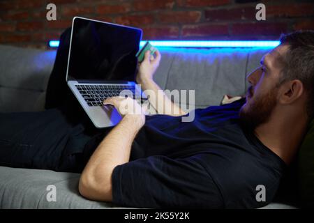 Fröhlicher, hübscher Kaukasusmann, der auf dem Sofa mit Laptop und Kreditkarte in der Hand im Wohnzimmer zu Hause liegt. Neonlicht. Online-Shopping und Stockfoto
