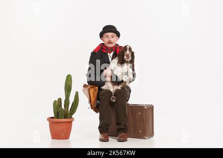 Portrait von stilvollen jungen Mann mit lustigen Ausdruck in Mantel und Hut sitzen auf Koffer mit Hund isoliert auf weißem Hintergrund Stockfoto