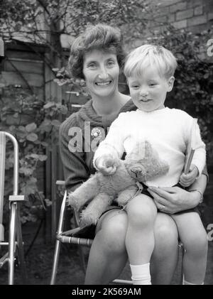 1960s, historisch, eine glückliche Mutter, die draußen auf einem metallenen Gartenstuhl sitzt, mit ihrem kleinen Sohn auf dem Schoß, der sein weiches Spielzeug, einen Teddybären, hält, England, Großbritannien. Stockfoto