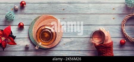 Wintertee, Panorama-Flachlage mit Glas-Teekannen, Glas Tee in der Hand auf verblassten Holzplanken. Weihnachtsdekor - Spiegelkugeln, Kugeln, Spielzeug Stockfoto