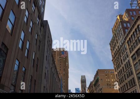 Wunderschöne Sicht von unten auf den blauen Himmel zwischen Wolkenkratzern mit US-Flagge an der Hauswand in New York. Stockfoto