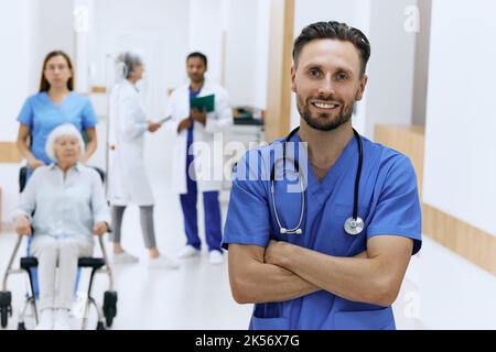 Hübscher Arzt mit Stethoskop, der vor der Krankenhauslobby mit Ärzten in weißen Mänteln, medizinischem Personal und Patienten steht. Arbeitstag Stockfoto