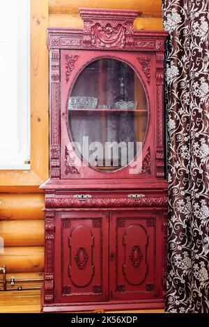 Alter alter, roter Holzschrank mit geschnitzten Dekorationen in der Ecke einer ländlichen Blockhütte zwischen Fenster und braunem Vorhang. Stockfoto