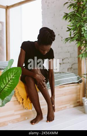 Schöne afro amerikanische Frau mit Dreadlocks sitzen auf dem Bett in dekorierten Studio und massieren Fuß von Hand. Schwarze Person mit Sportverletzung, Muskel Stockfoto