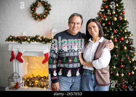 Glückliches multiethnisches Paar lächelt der Kamera im Wohnzimmer mit dekorierten Kamin und weihnachtsbaum, Stockbild Stockfoto