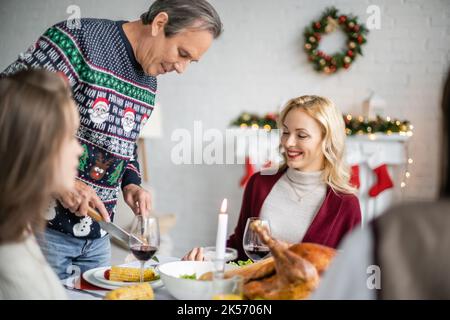 Älterer Mann schneidet gegrillten Mais und lächelt in der Nähe der Familie beim weihnachtsessen, Stockbild Stockfoto