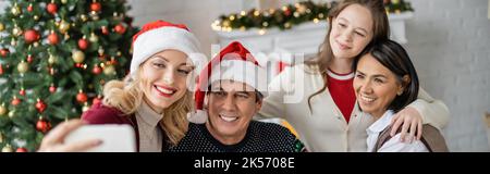 Glückliche Frau im weihnachtsmann hat Selfie mit Tochter und multiethnischen Eltern in der Nähe verschwommener weihnachtsbaum, Banner, Stockbild Stockfoto
