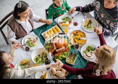 Draufsicht auf die multiethnische Familie, die sich die Hände hält und in der Nähe einer köstlichen Mahlzeit betet, bevor man sich beim Abendessen für Danksagebete begibt, Stockbild Stockfoto