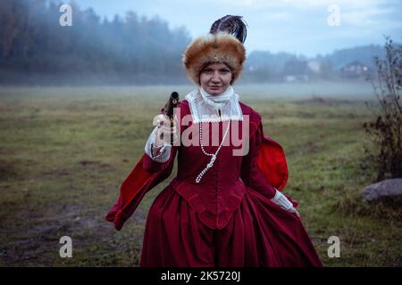 Eine Frau in einem roten Kleid aus dem 17.. Jahrhundert mit einer Duellpistole zielt auf die Kamera Stockfoto