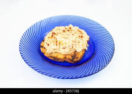 Humuspaste mit Paprikapulver auf einer gerösteten Brotscheibe, auf einer blauen Glasplatte, isoliert auf weißem Grund. Seitenansicht Nahaufnahme. Stockfoto