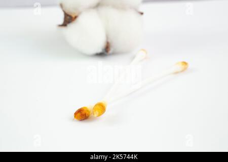Wattestäbchen, die nach der Ohrenreinigung mit einer im Hintergrund entkochten Baumwollblume mit Cerumen verschmutzt sind Stockfoto