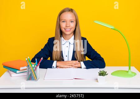 Foto von optimistischen smart clever Schulmädchen mit geraden Frisur tragen blauen Blazer sitzen am Schreibtisch isoliert auf gelbem Hintergrund Stockfoto