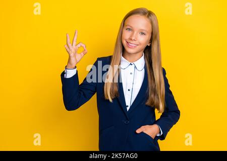 Foto von positiven fröhlichen Schulmädchen mit geraden Frisur tragen blauen Blazer zeigt okey genehmigen isoliert auf gelbem Hintergrund Stockfoto