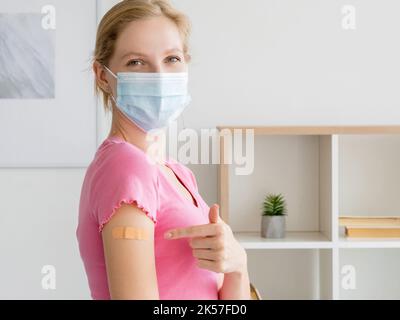 Schutzimpfung. Glückliche Frau. Bleib sicher. Lächelnde Dame in einer Medizinmaske, die auf Injektionsputz im hellen Innenraum zeigt. Stockfoto