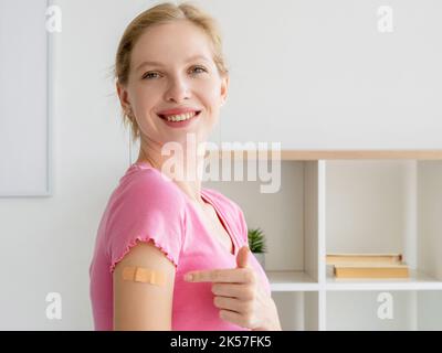 Covid-Schutz. Glückliche Patientin. Impfergebnis. Zufriedene Frau zeigt auf Gips in der Schulter nach der Injektion in hellen Raum Innenraum. Stockfoto