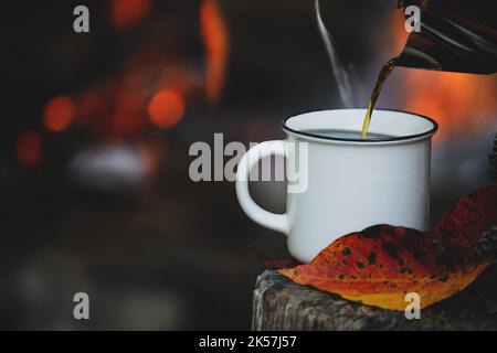 Wasserkocher gießt heißen, dampfenden Kaffee in eine weiße emaillierte Tasse, die auf einem alten Baumstamm mit Herbstblättern sitzt. Selektiver Fokus auf Becher mit unscharfem Hintergrund. Stockfoto