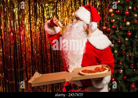 weihnachtsmann in Kostüm hält köstliche Pizza zum Mitnehmen in der Nähe von weihnachtsbaum und Lametta, Stock Bild Stockfoto