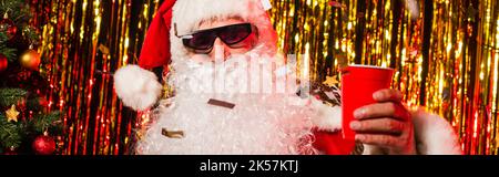 weihnachtsmann in Sonnenbrille hält Plastikbecher unter Konfetti in der Nähe Lametta, Banner, Stock Bild Stockfoto