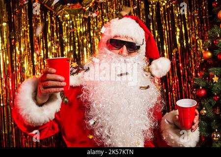 weihnachtsmann in einer Sonnenbrille mit Plastikbechern unter Konfetti in der Nähe des weihnachtsbaums, Stockbild Stockfoto