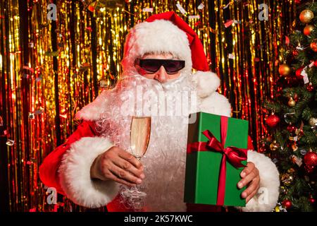 weihnachtsmann in einer Sonnenbrille mit Sektglas und Geschenk in der Nähe von weihnachtsbaum und Lametta, Stockbild Stockfoto