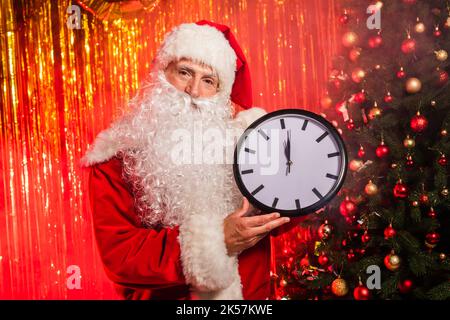 weihnachtsmann in Kostüm hält Uhr in der Nähe von weihnachtsbaum und Lametta, Stock Bild Stockfoto