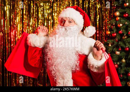 weihnachtsmann in Kostüm hält Einkaufstaschen in der Nähe von weihnachtsbaum und Lametta, Stock Bild Stockfoto