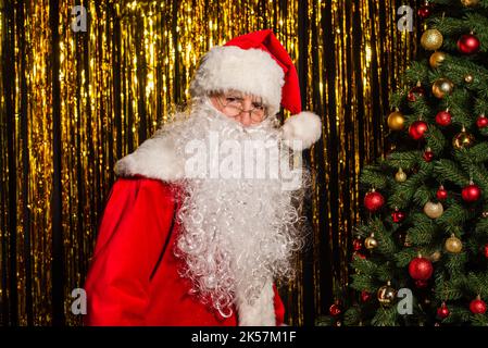 weihnachtsmann in Kostüm und Brille Blick auf Kamera in der Nähe von weihnachtsbaum und goldenes Lametta, Stock Bild Stockfoto