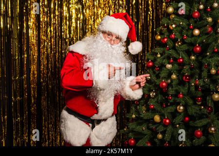 weihnachtsmann zeigt mit den Fingern auf geschmückten weihnachtsbaum in der Nähe von Lametta, Stockbild Stockfoto