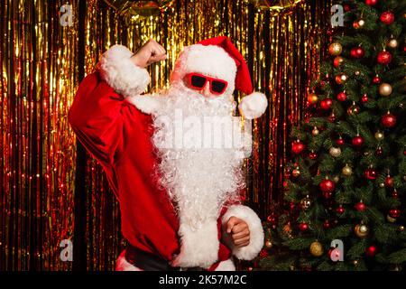 weihnachtsmann in Sonnenbrillen und Kostüm tanzen in der Nähe von weihnachtsbaum und Lametta, Stock Bild Stockfoto