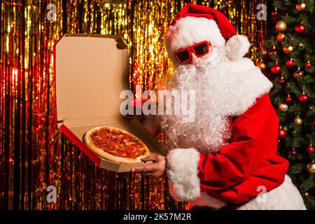weihnachtsmann in Sonnenbrille mit Pizza zum Mitnehmen in der Nähe von weihnachtsbaum und Lametta, Stockbild Stockfoto
